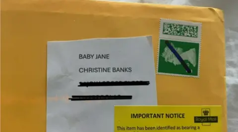 The envelope of a letter sent to Christine Banks. It is a yellow envelope with the words BABY JANE, CHRISTINE BANKS written on it. It also has a green stamp that a line has been drawn through, and a yellow Royal Mail warning that the letter is bearing a counterfeit stamp.