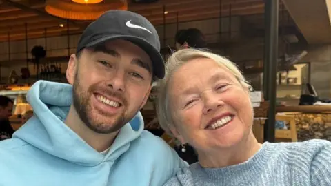 Courtesy of Quirke family for Alzheimer's Research UK (L-R) A son and mum. The son is wearing a blue hoodie and a black sun hat. The mum is wearing a grey/blue sweater. They are both smiling at the camera and appear to be sitting in a cafe. 