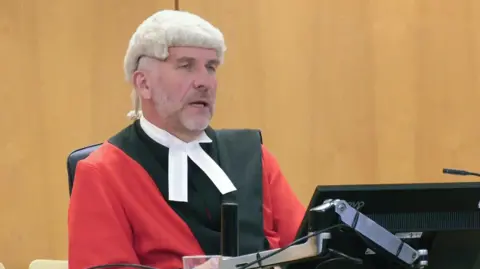 PA Media A judge wearing a cap and gown while sat in a courtroom delivering a sentencing in front of a monitor.