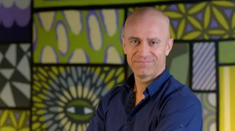 Supplied A man with a bald head and blue eyes smiles at the camera in front of a patterned wall.