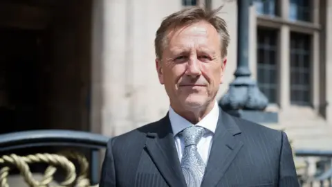 Staffordshire County Council Martin Murray, a man wearing a dark grey suit is standing outside Stafford CountyBuildings. He is also wearing a white shirt and pale-coloured tie.
