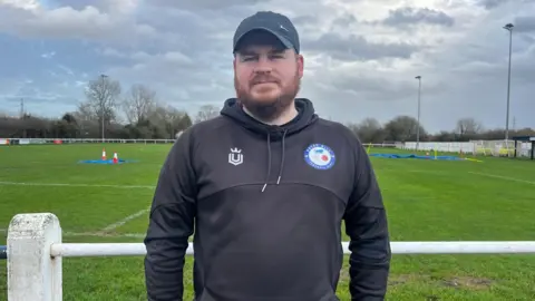 Picture of Aaron standing in front of the football pitch. He's wearing a black hoodie and cap and has a thick beard.