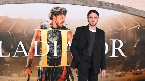 Getty Images Paul Mescal wears a black suit and has short brown hair. He stands in front of a billboard which says Gladiator II and features a photo of him wearing a metal breast plate. 