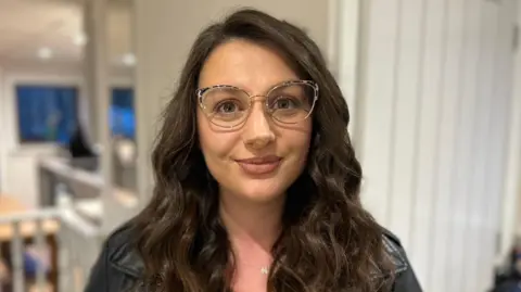 Woman with long wavy dark brown hair smiling at the camera. She is wearing glasses and a black leather jacket.