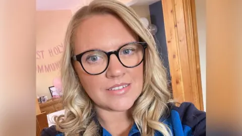 Leanne Flanagan Women with blonde long hair looking at camera. She is wearing black glasses and a blue jumper.