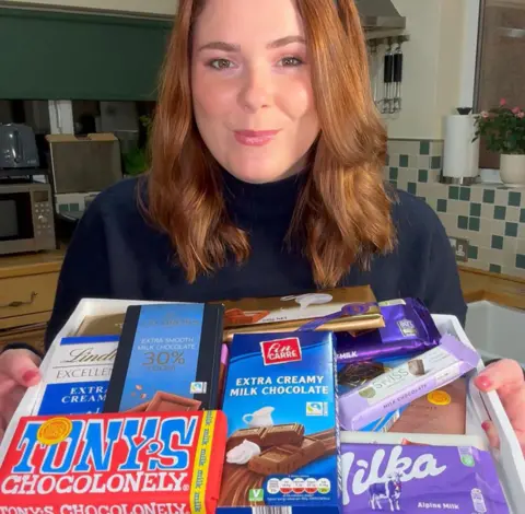 Becca Amy Stock Becca Amy Stock is smiling and looking at the camera. In front of her she is holding a tray of milk chocolate bars, all different brands.