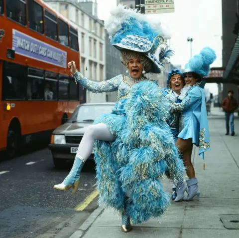 A man dressed up in a lavish blue costume dancing on the pavement with a bus in the background