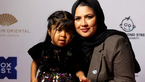 PA Media Fahima kneels next to Zarwa as they pose together on a red carpet in front of a charity branding backdrop.