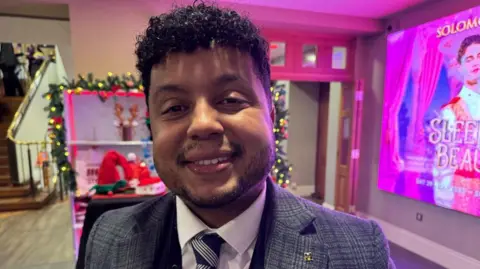 A mid shot of a man in a grey suit, white shirt and blue and striped tie. He has short dark curly hair and is standing in a foyer that is decorated with Christmas decorations. He is smiling. 