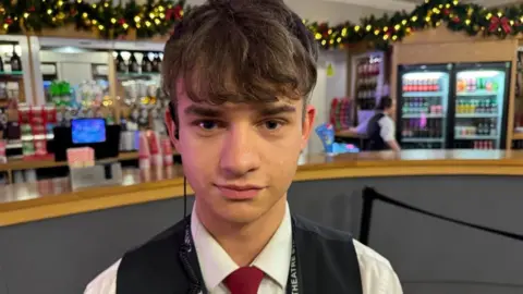 A young man who is standing in front of  bar at a theatre. He is wearing a white shirt and a black waistcoat as well as a red tie. The bar in the background is decorated with Christmas garland and lights. 