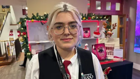 A young woman with blonde hair that is tied back with a centre parting. She is wearing a white shirt, black waistcoat and red tie. She is smiling and standing a theatre foyer that is decorated with Christmas decorations. 