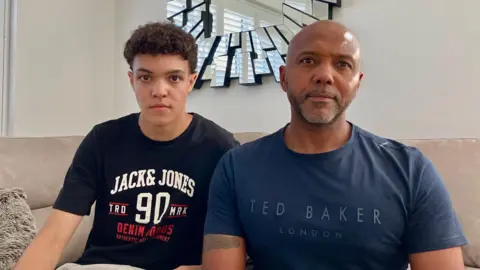 The same young man as before sitting next to an older man who is bald and has a short greying goatee and is wearing a blue t-shirt