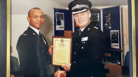 Rod Rose Two police officers in uniform. They are shaking hands and the man on the right is handing the man on the left a certificate in a frame.