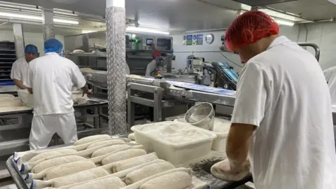 A baker folds dough into a loaf. Next to him are around a dozen loaves, already formed.