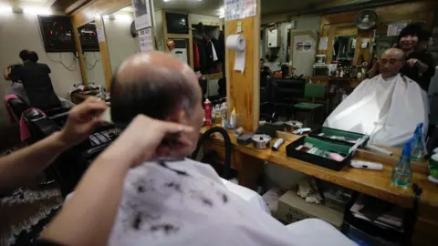 Getty Images Backview of an old man, who's balding, getting a haircut at the barbers. You can see his face through the mirror in front of him.