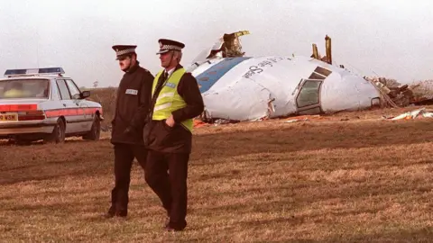 Getty Images An archive image of the nose cone section of the plane which crashed in a Lockerbie field shows two policemen walking in front of the wreckage, a police car situated at the left of the plane.