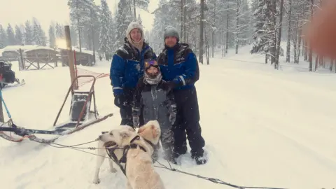 Rich Stephenson-Evans Two men in thick blue and black coats are standing with a boy, who is also wearing a thick coat. They are in Lapland. Standing next to them are two sleigh dogs.