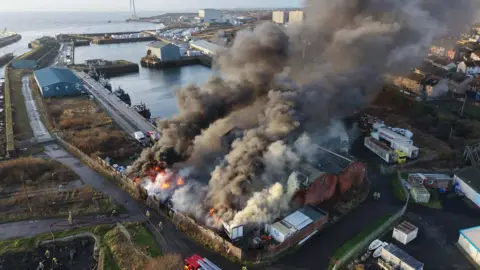 Fife Jammer Locations Wide aerial shot of Methil Harbour with a large fire producing dense black smoke and flames near industrial buildings. Fire engines and crews surround the site, with the harbour, boats, and a wind turbine visible in the distance.