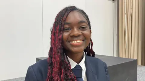 Kate McGough / BBC A teenage girl with long red hair is smiling at the camera. She's wearing a blue school tie, white shirt and blue jacket and sitting on a school stage. 