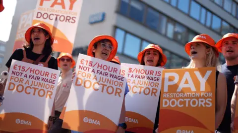 PA Media A group of junior (now called resident) doctors holding placards outside a hospital in London last year. They are holding signs saying "£15 an hour is not a fair wage for a junior doctor" and wearing orange hats with the BMA logo on them. They look like they are singing or chanting.