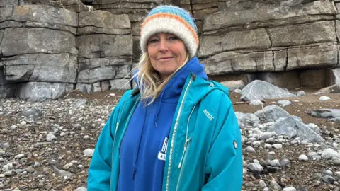A woman wearing a white, orange, turquoise and blue woolly hat, a blue hooded top and a turquoise coat is smiling while standing on a beach. 