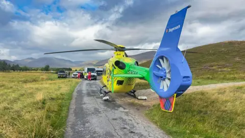 SCAA A rescue helicopter in bright yellow and green with a blue tail is on a hillside with ambulances and cars in front of it