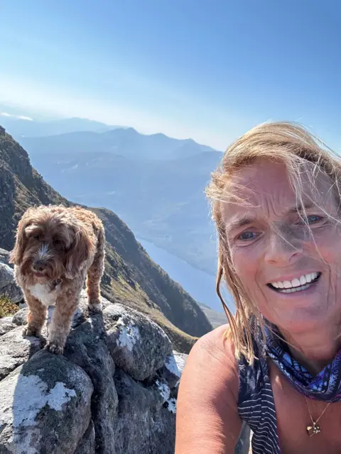 Mary-Jane Parker A selfie of Mary-Jane who has blonde hair. She is with her dog at the top of a Munro with a valley in the background