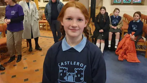 Elsie smiling at the camera. She has ginger hair and is wearing her school uniform which is navy and light blue.