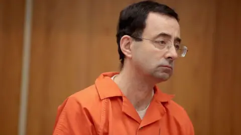 Getty Images Larry Nassar from the shoulders up, wearing square framed glasses with wire rims and orange jumpsuit, looks off to his left