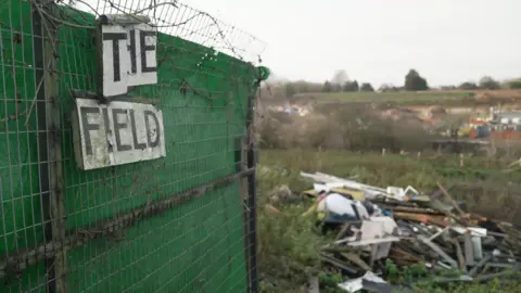 Fiona Irving / BBC An open green gate with a sign reading "The Field" on it. Behind the sign, an overgrown field with fly-tipped rubbish.