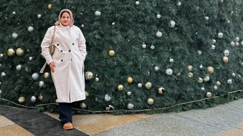 North West Migrants Forum Farida Hassan is standing in front of a huge Christmas tree - only the bottom is showing. She is wearing a beige jacket and a headscarf. 