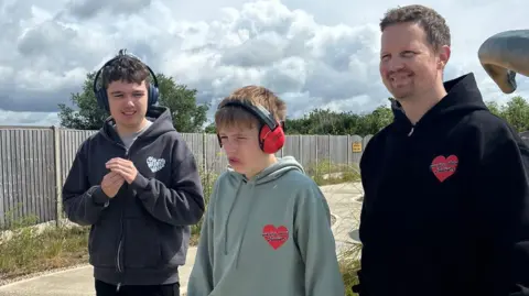James Hunt Jude wearing ear defenders and a grey hoodie next to Tommy in ear defenders and in a green hoodie and their Dad James who is wearing a black hoodie