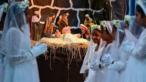 Getty Images Girls stand alongside a Christmas nativity scene depicting the birth of Jesus during a Christmas mass at St Mary's Church in the village of Uswetakeiyawa, Sri Lanka on 25 December 2025. 
