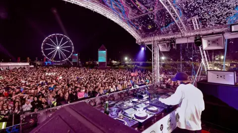Pavilion Festival Ewan McVicar performing on stage at a previous Pavilion festival - thousands of people are watching and a big wheel is in the distance. The view is from behind Ewan, who is is DJing at his decks.  