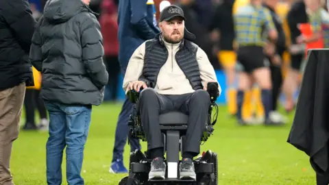 PA Ed Slater, who is using a motorised wheelchair, makes his way across a rugby pitch, where people are milling around. Ed is wearing a black cap with a "4ED" logo, a light coloured three quarter zip jumper, a black bodywarmer, black trousers, and grey trainers.