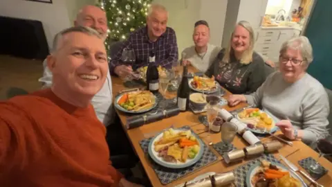 One Day More Aboard Six people, four men and two women smiling for a selfie at a table. They are eating Christmas lunch with crackers between them. There is a lit up Christmas tree behind them