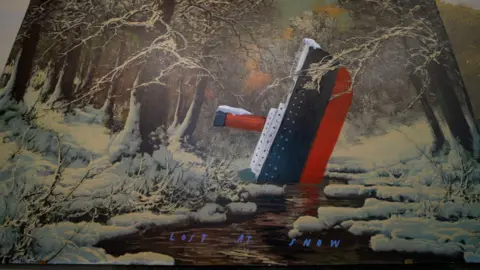 A close up of an artwork. It depicts a boat which resembles the Titanic capsized in a stream in a forest. The trees, boat and landscape are covered in snow. Lost At Snow is written in blue at the bottom.