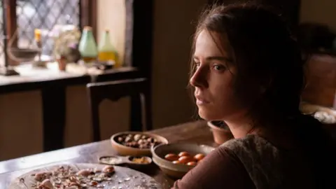 Focus Features Jessie Buckley as Agnes in Hamnet, dark hair and a pensive expression, sitting at a table with food on it