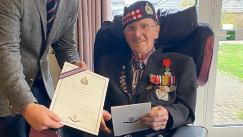 KOSB Douglas Baldwin with his birthday gift from the KOSB Association. He is smiling as he sites in a chair. He is wearing a beret and jacket with medals on the lapel. He is holding a card and someone is leaning in to the picture to show a special birthday certificate to the camera.