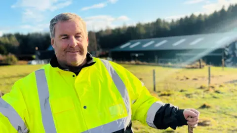 Mike Duxbury smiling at camera, he is wearing a bright yellow safety jacket, there is a farm building in the distance behind him.