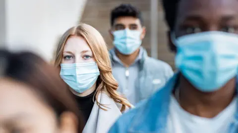 Getty Images People wearing blue surgical masks. The photo is focusses on a woman with a black jumper and grey coat on. She has long fair hair.