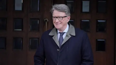 PA Media Mandelson smiling. He is wearing black framed glasses and a navy coat with white shirt and blue tie. He is standing in front of a wooden door with a gold handle.