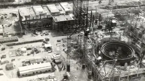 Nuclear Restoration Services (NRS) A black and white aerial photo of Wylfa under construction. Large piles of scaffolding are visible, surrounding a variety of industrial buildings.
