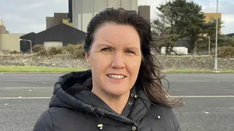 Ffion Morris stood in front of Wylfa. It is a head shot of her. She has a black puffa coat and a is looking at the camera. Her dark hair is a little windswept. 