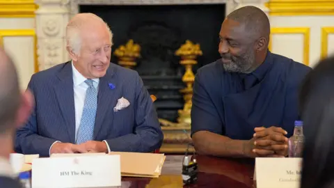 PA Media King Charles III, wearing a blue pinstripe suit and a blue patterned tie, looks across and grins as he sits next to Idris Elba, who smiles back. Elba is wearing a contemporary African-style suit in navy. The pair are sitting with others at a meeting table in St James's Palace, with an ornate fireplace behind them and gilded panelling visible in the background.