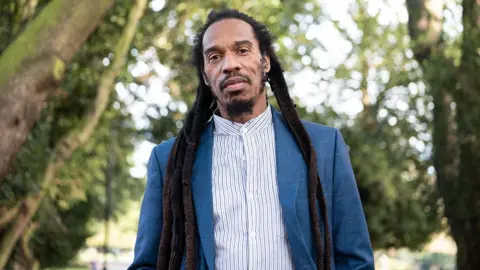 The poet Benjamin Zephaniah is pictured in a park wearing a white and blue striped shirt and a blue jacket. 