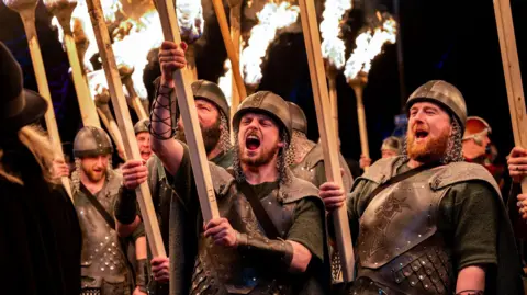 Ian Georgeson Men dressed as vikings wearing dark leather outfits and armour and metal headgear with chainmail hold up flaming torches.