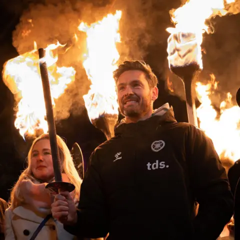 Ian Georgeson Craig Gordon wearing a jacket with the Heart of Midlothian badge on it holding a flaming torch and smiling just off camera.