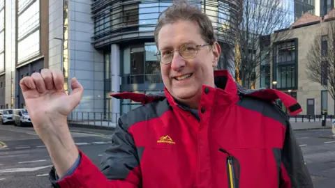 Jonathan Burns, wearing a red waterproof coat with grey sleeves, holding his hand up to the camera with a very small scar - no more than a centimetre - visible on his wrist. He has brown hair and round glasses. He is standing in Bedford Street, behind him is a modern high-rise building of  glass and steel.