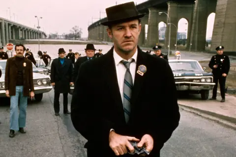 Getty Images Gene Hackman filmed in a scene from The French Connection. He wearing a dark overcoat, white shirt, and striped tie and stands in the foreground holding a small object in both hands. Hackman also wears a dark fedora-style hat and has a badge pinned to the coat. Behind him, several uniformed police officers and other individuals are visible near two vintage police cars. The scene takes place outdoors on a road beside large concrete bridge structures, with an overcast sky in the background.